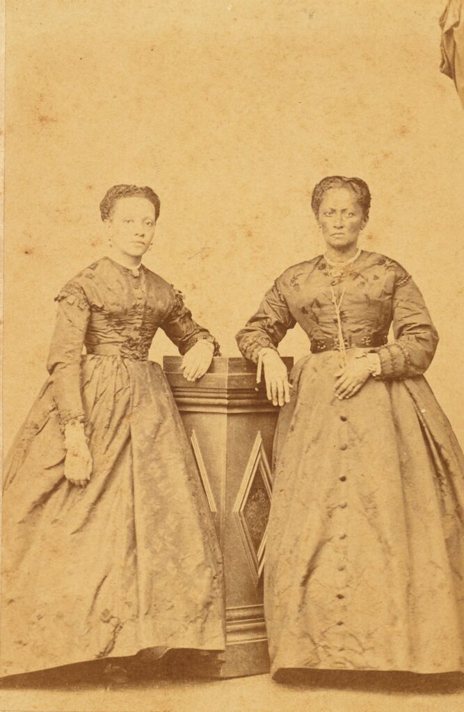 Duas mulheres negras posam em estúdio com roupas e adornos elegantes.